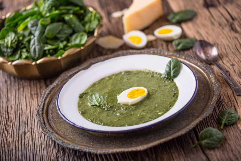 Green soup in a white bowl on a table with spinach eggs and cheese in the background