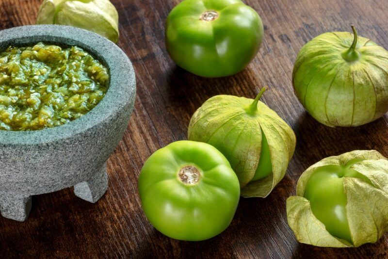 A wooden table with green tomatillos and a small container of salsa verde