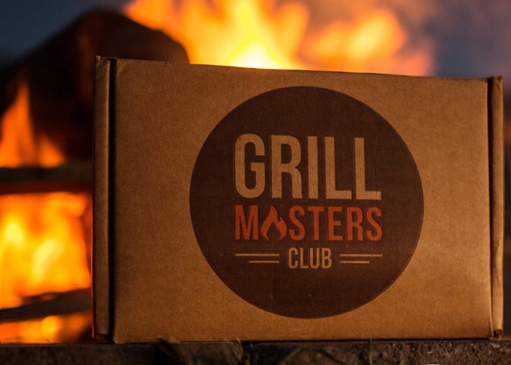 Cardboard Grill Masters Club box sitting on a tree stump.  Open bonfire in the background.  Box has a brown circle that stated Grill Masters Club in the Middle