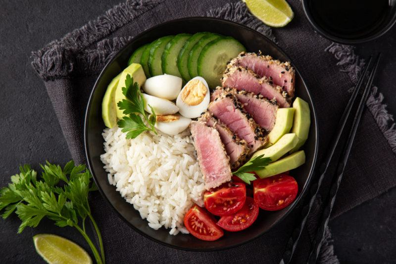 A black bowl with sliced tuna steaks, rice and greens