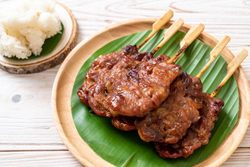 A bamboo plate with a banana leaf and grilled pork on sticks, next to some sticky rice