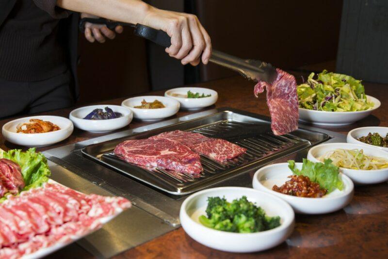 Someone cooking steak on an indoor bowl, with many bowls of vegetables surrounding it