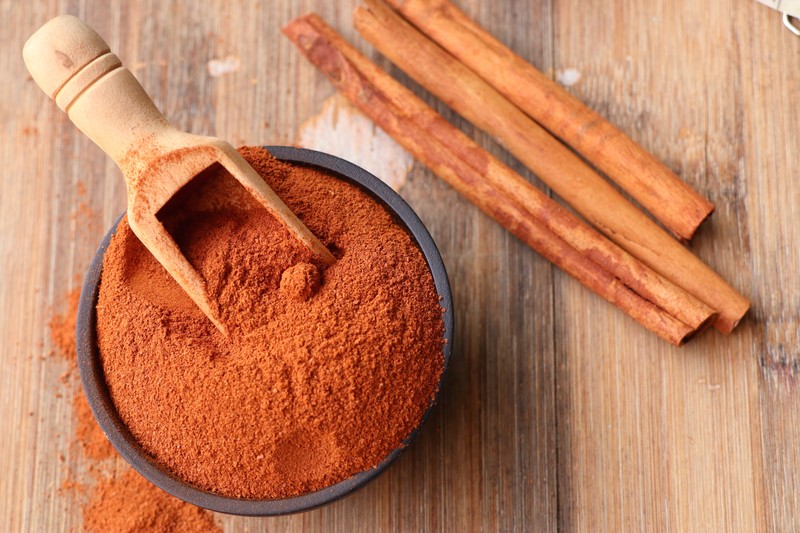 top view image of a black bowl full of ground cinnamon with wooden scoop, on top of a wooden surface with cinnamon sticks and loose cinnamon powder