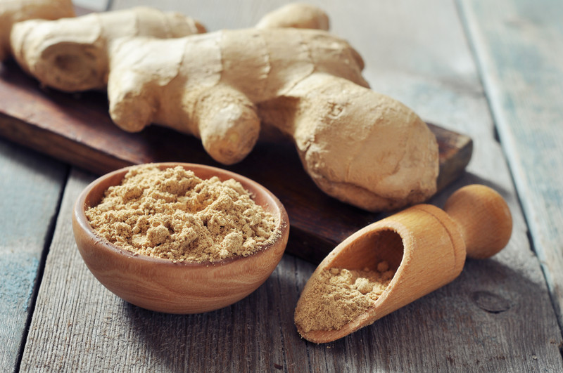 A wooden bowl and scoop with ground ginger on a wooden table with fresh ginger at the back resting on a wood plank.