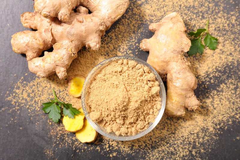 top view image of a glass bowl full of ground ginger, with fresh ginger roots, sliced ginger, and coriander leaves beside it