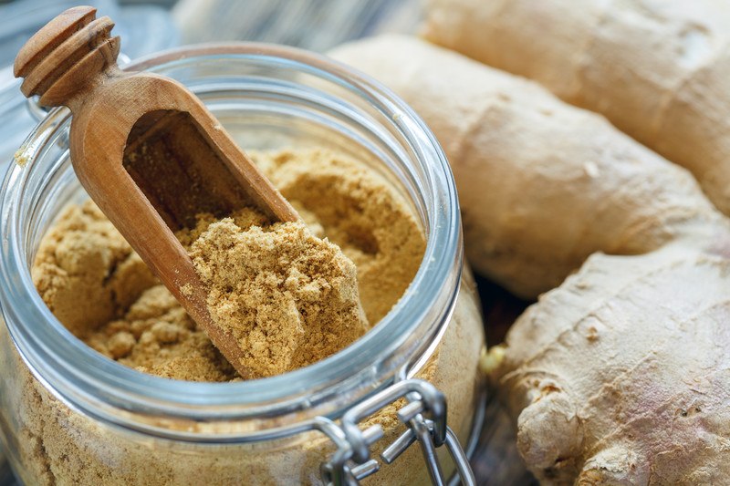 Jar of ground ginger in a mason jar with a small wooden scoop, with fresh ginger on the side.