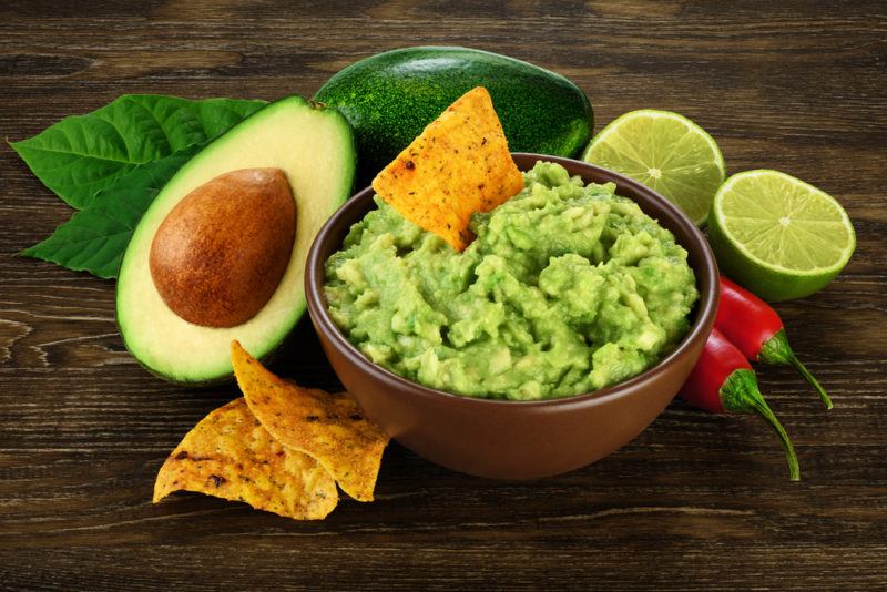 A brown bowl with guacamole, next to tortilla chips, avocados, limes and chili