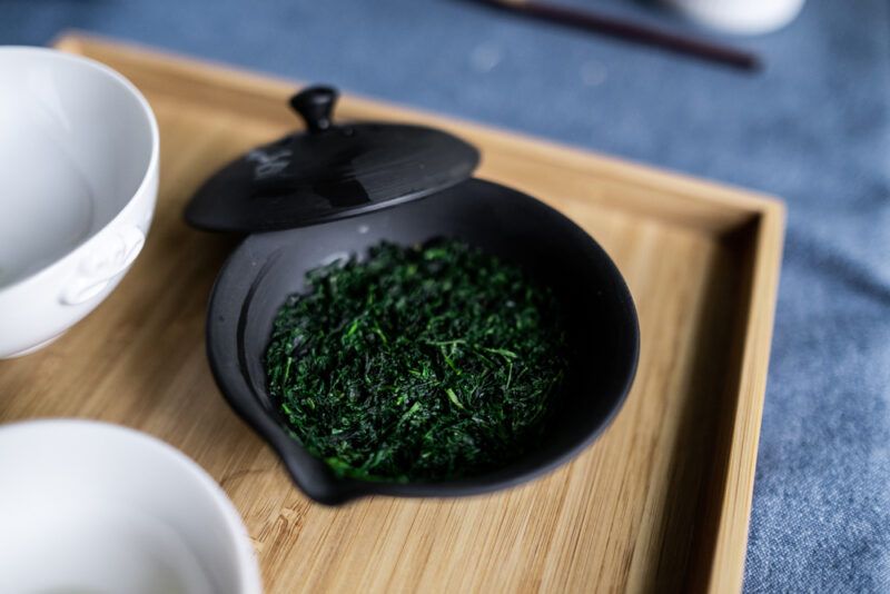 on a black ceramic dish are loose Gyokuro tea resting on a wooden tray with white ceramic tea cups beside it