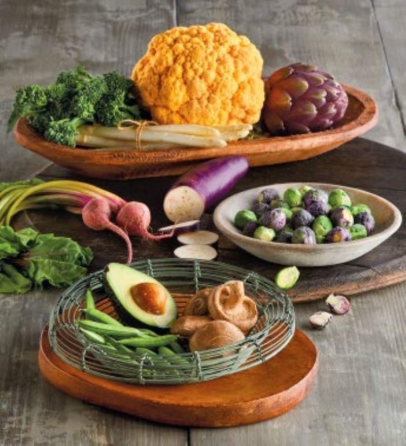 GRAY WOODEN TABLE WITH WOODEN BOWLS WITH COLORFUL VEGETABLES SUCH AS, ORANGE CAULIFLOWER, PURPLE ARTICHOKE, WHITE ASPARAGUS, AVOCADO, SHIITAKE MUSHROOM, GREEN AND PURPLE BRUSSELS SPROUTS, AND BEETS