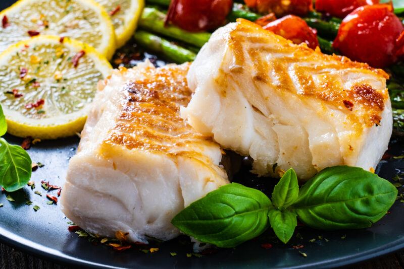 Fish,Dish,-,Fried,Cod,Fillet,With,Asparagus,On,Wooden | Food For Net This photo shows two pieces of cooked halibut on a dark plate near lemon slices and mint leaves.