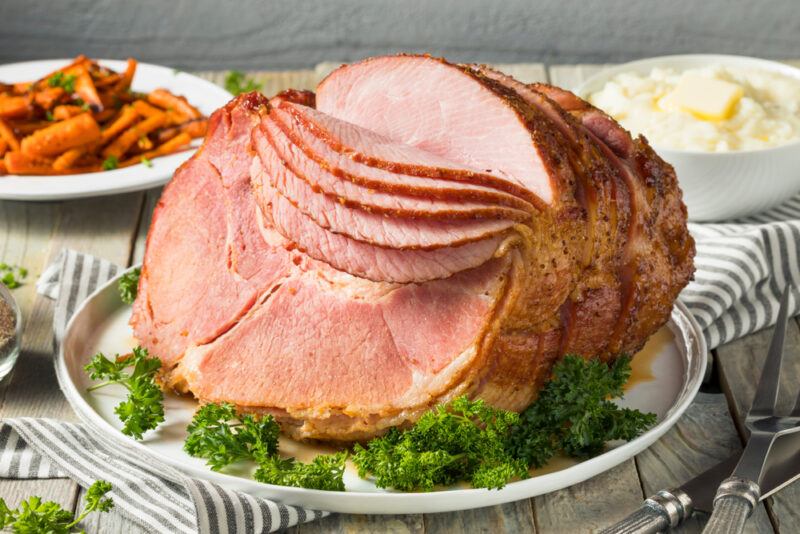 Homemade,Glazed,Easter,Spiral,Cut,Ham,With,Carrots,And,Potatoes | Food For Net This photo shows a cooked, sliced ham on a white plate with parsley sprigs, near a bowl of mashed potatoes and a bowl of braised carrots.