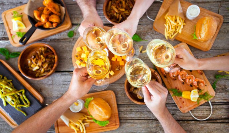 Various hands holding wine glasses and toasting on top of a table that is covered with Thai dishes