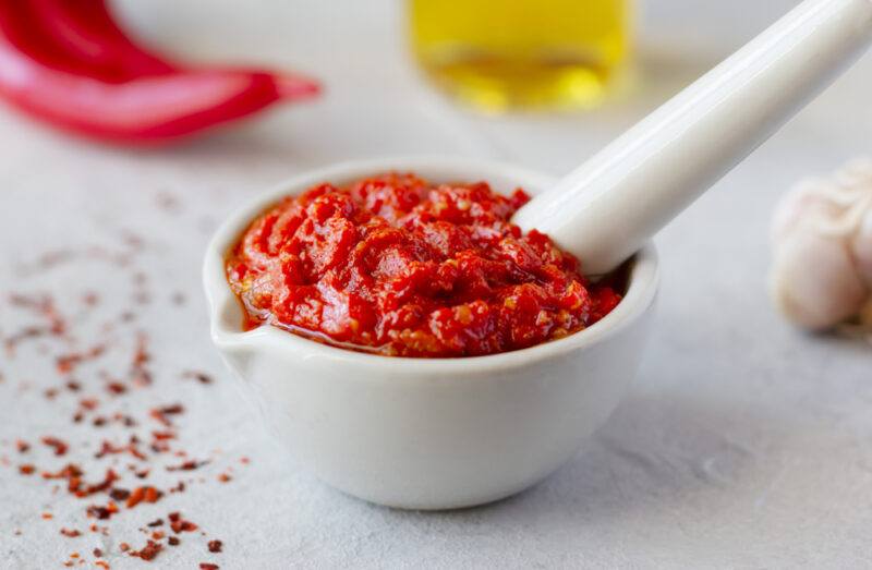 on a white surface is a white mortar and pestle with Harissa, around it is loose chili flakes, fresh peppers, jar of olive oil, and garlic cloves around it