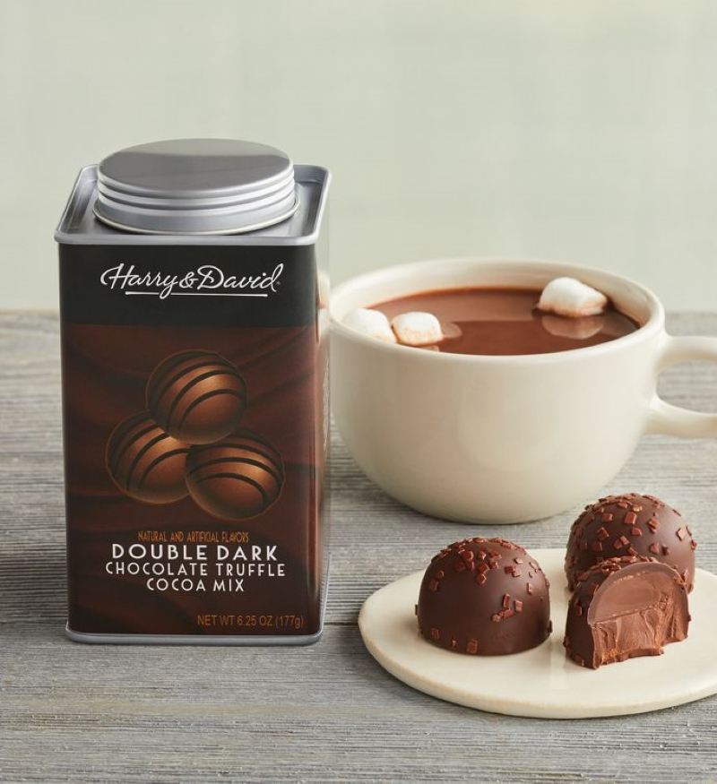 Harry and David canister of double dark chocolate truffle hot cocoa next to the canister is a small plate with two whole and one half truffle and behind that is a white much with hot cocoa and three mini marshmallows floating on top