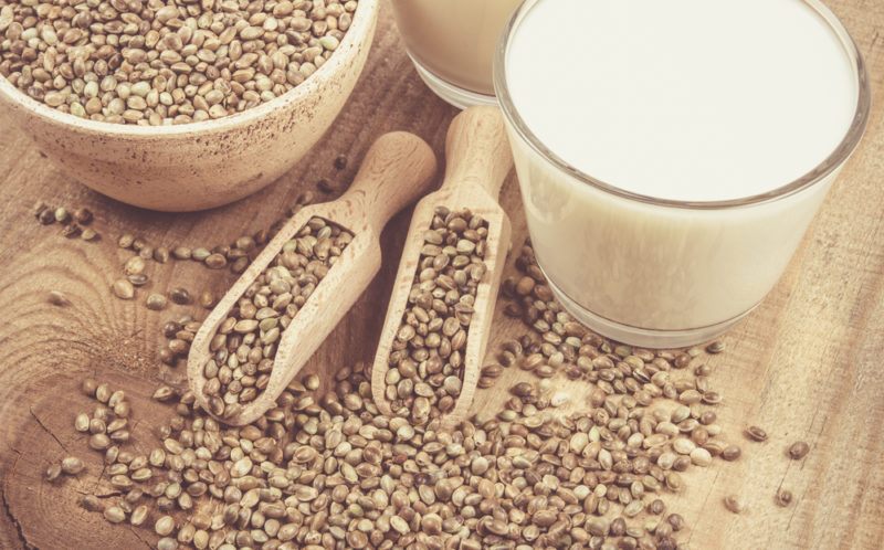 A bowl of hemp seeds, some spoons of hemp seeds and a glass jar of hemp milk