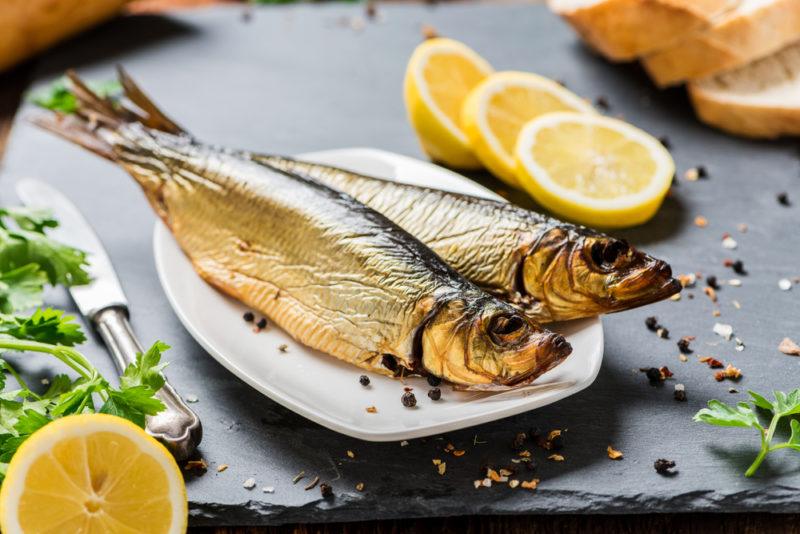 A white plate with two cooked herrings, with slices of lemon and cracked pepper on a table