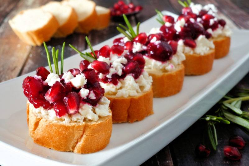 An oblong white plate with five pieces of crostini that have been topped with berries and cheese to make a holiday appetizer