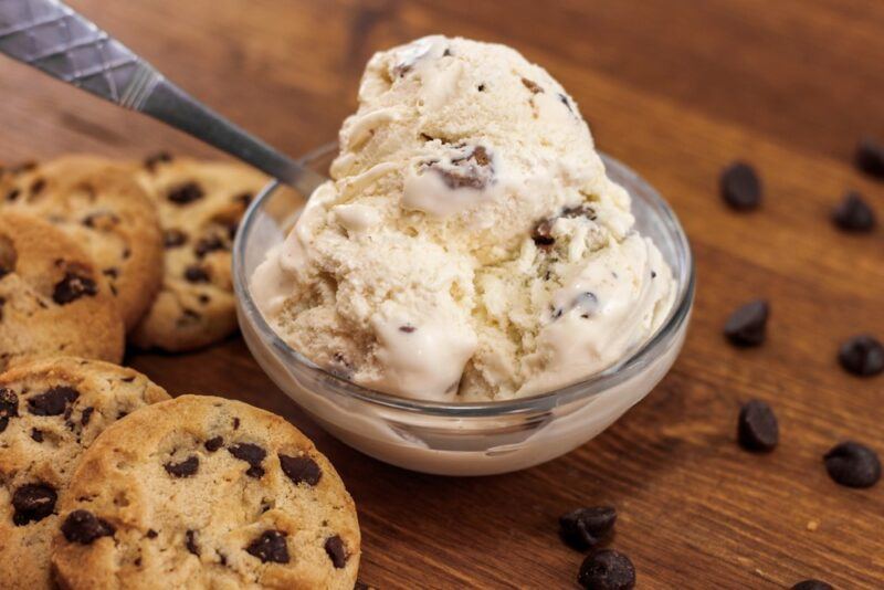 A glass dish of cookie dough ice cream with a spoon, next to some baked cookies