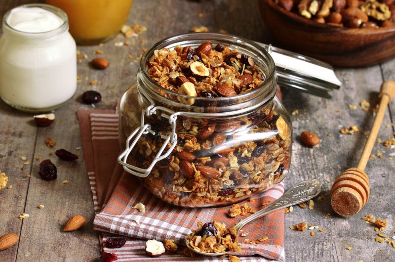 A glass jar filled with homemade granola, next to a spoon of granola and some yogurt