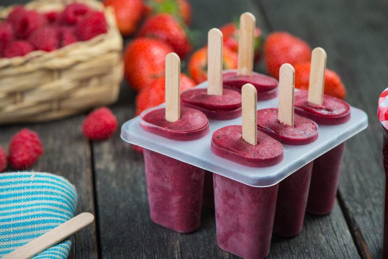 Six berry popsicles in molds on a table, with berries in the background