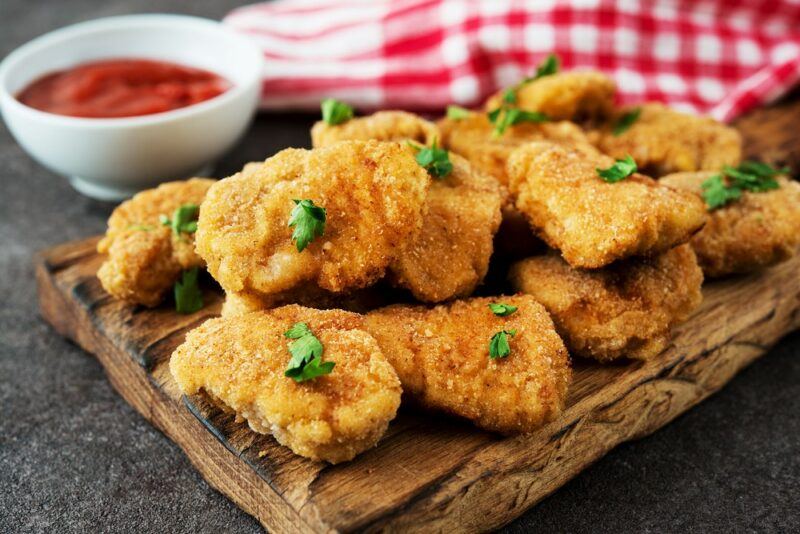 A wooden board with homemade chicken nuggets and a small white bowl containing tomato sauce