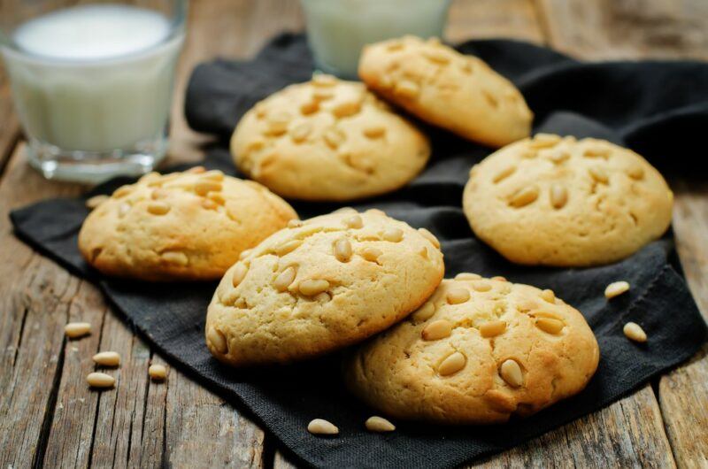 Six freshly made cookies with pine nuts on top of them, in front of a glass of milk