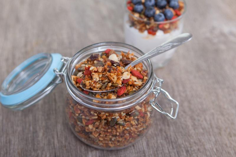 A glass jar with a lid containing homemade granola, with a parfait in the background