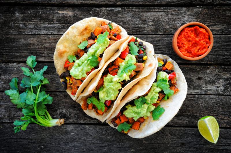 Three homemade tacos on a dark wooden table with some greens, a lime, and seasoning on the side