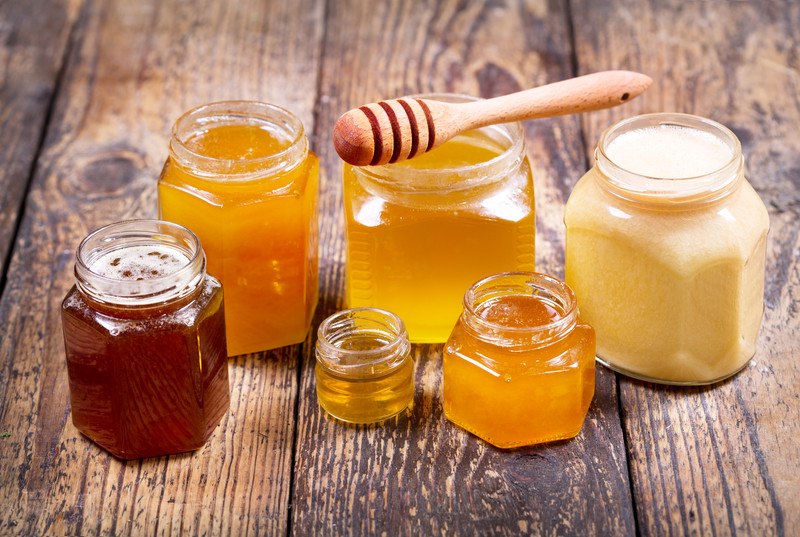 This photo shows six jars of honey in varying sizes and shades sitting on a wooden table.