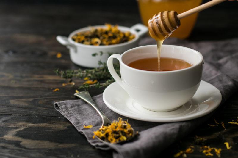 Honey being drizzled from a wooden stick into a mug of herbal tea