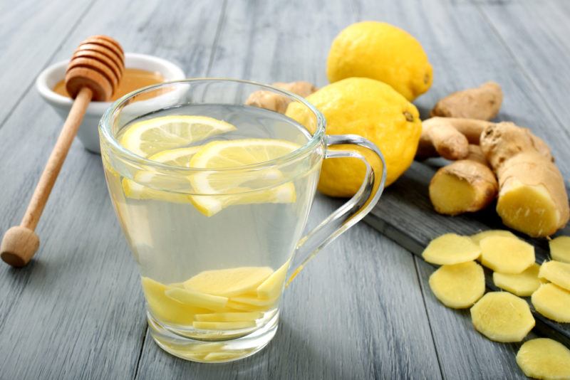 A glass mug containing hot lemon water with lemon, ginger and honey in the background