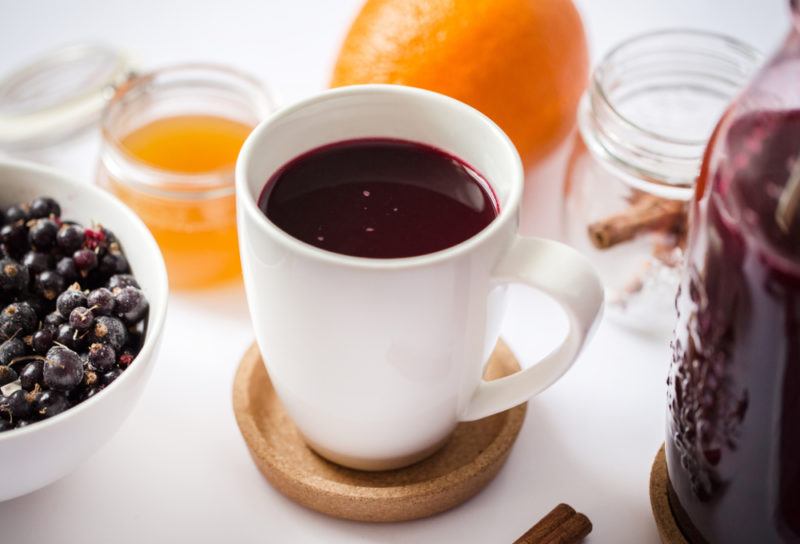 A white mug containing hot blackcurrant drink with blackcurrants and lemon in the background