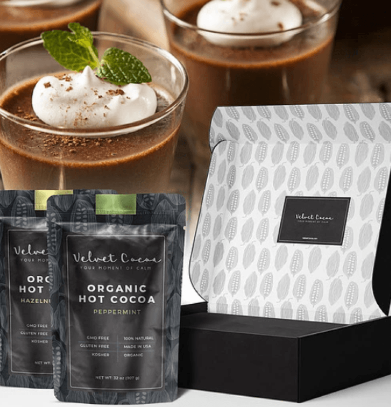 Velvet Cocoa hot chocolate box open in the lower right corner with two- two pound bags of hot cocoa sitting in front,  In the background are glass mugs of hot chocolate topped with whipped cream and garnished with grated chocolate and mint leaves