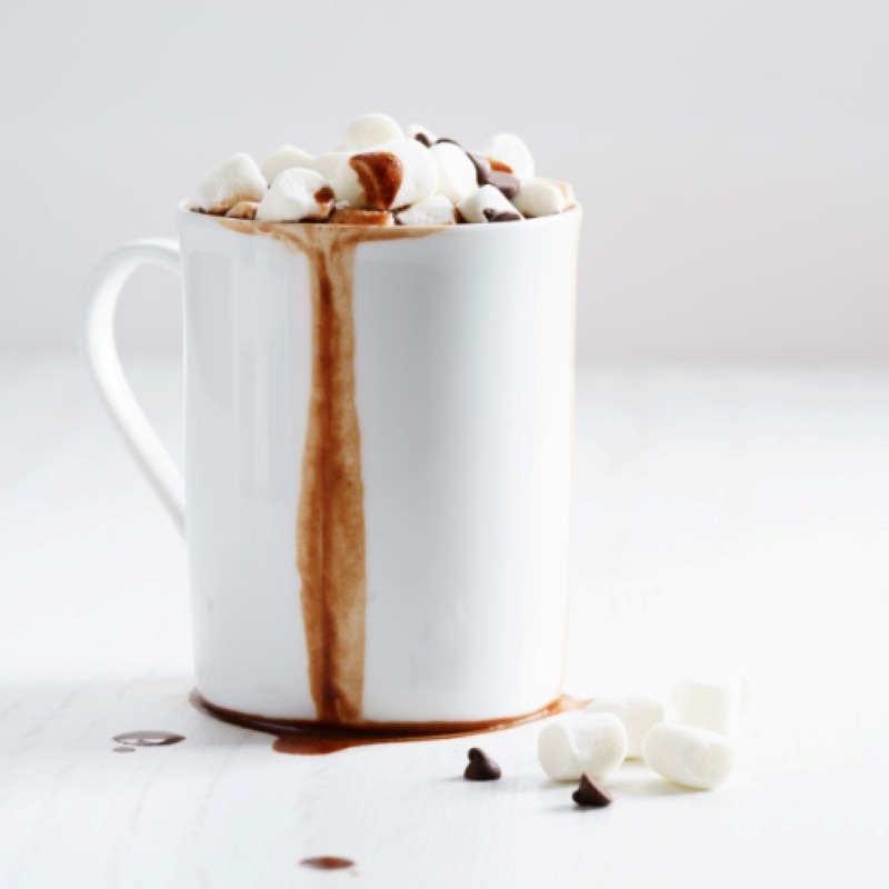 White mug overflowing with hot chocolate topped with mini marshmallows and mini marshmallows and mini chocolate chips scattered in front of the mug on the table