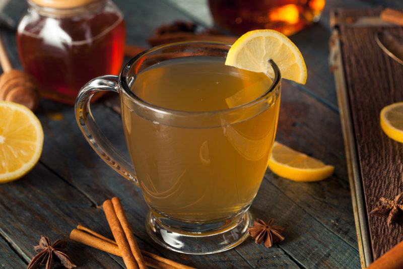 A hot toddy in a glass mug with cinnamon sticks, lemon and other ingredients on the table