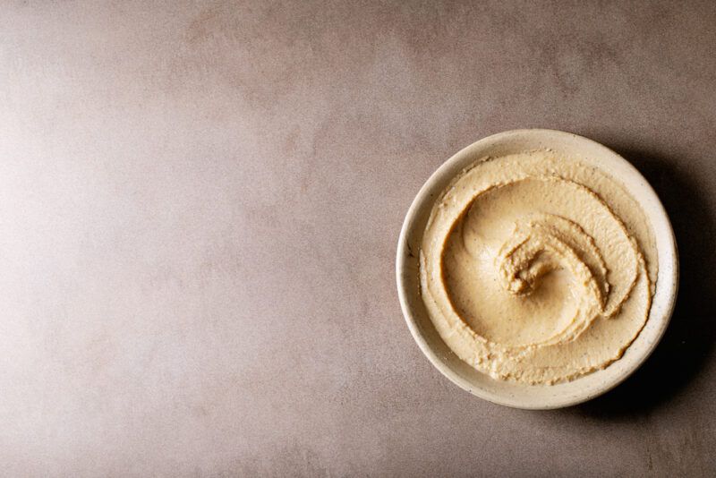 A bowl of hummus rests on a beige surface.