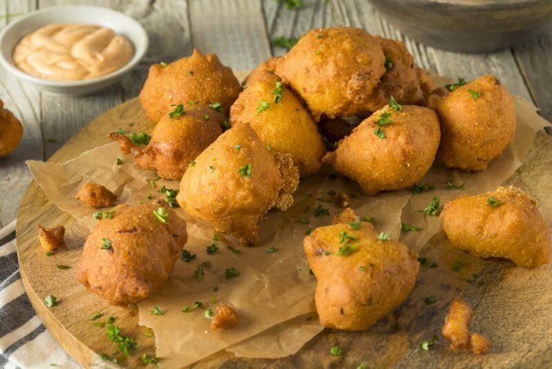 A collection of hush puppies on a wooden board, next to a small bowl of dip