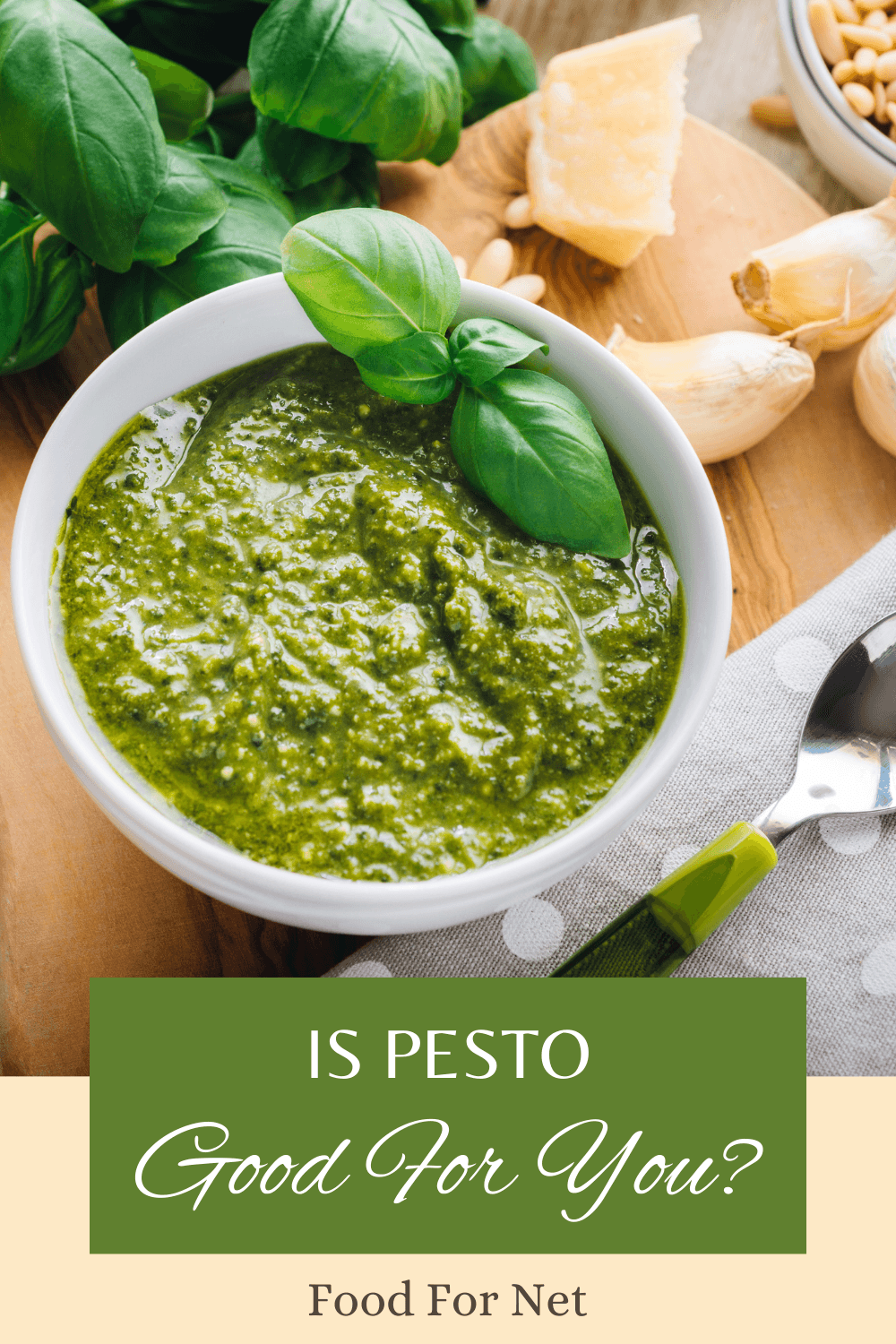 A white bowl of pesto with a basil leaf, with garlic, pine nuts, basil, and parmesan in the background, highlighting the question of whether pesto is good for you