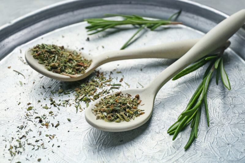 a silver platter with a couple of wooden spoons full of Italian seasoning, around them are a loose dried herbs and a couple of fresh herbs at the back