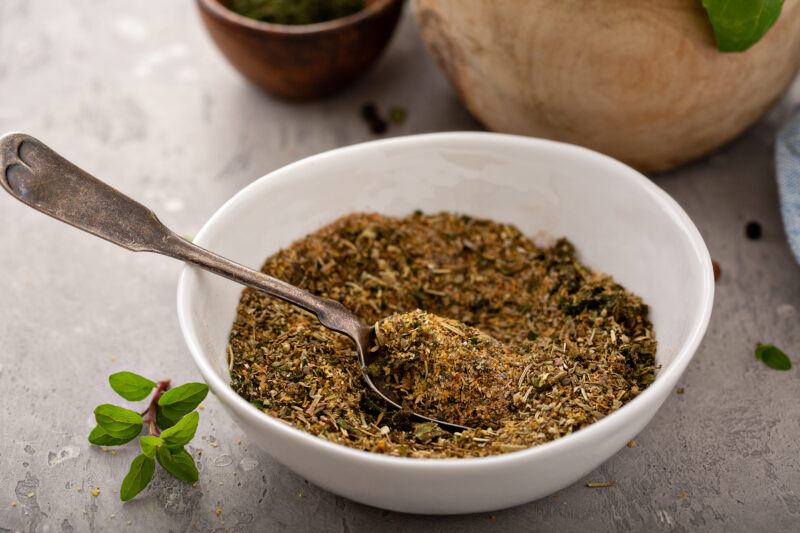 on a concrete surface is a a white bowl with Italian seasoning with a silver spoon shoved in it, around it is a small sprig of herb and a few loose peppercorns