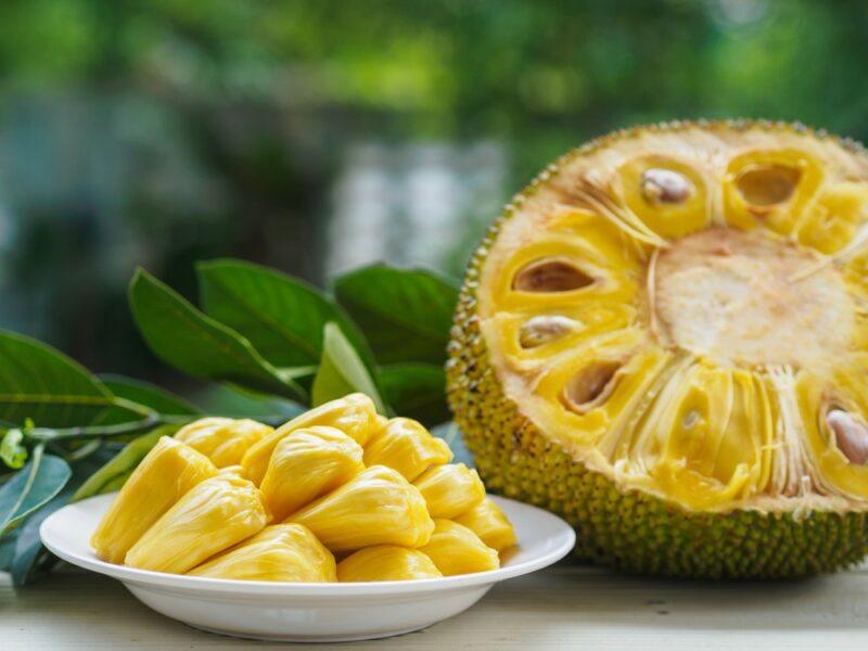 Half a jackfruit on a table, next to a white plate of jackfruit flesh