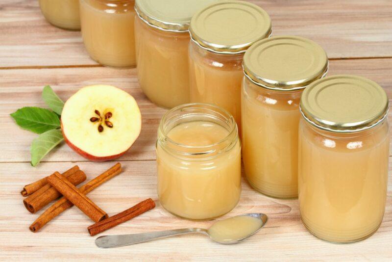 A line of applesauce jars with lids, next to one open one, some cinnamon, a spoon, and half an apple