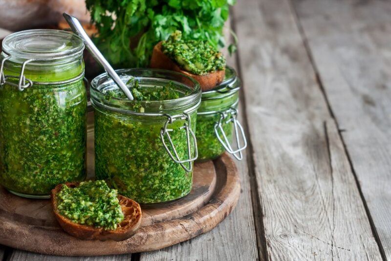 A wooden board with two jars of cilantro pesto and a spoon sticking out of one of them