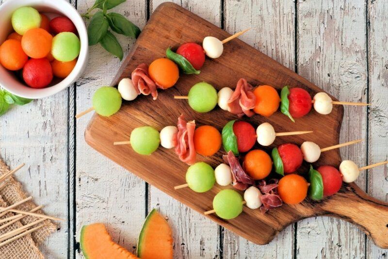 A wooden board with a selection of fruit kebabs, next to a small bowl of fruit balls