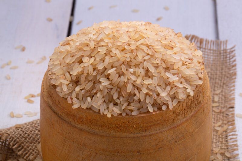 a white wooden surface with a wooden bowl of raw Kerala matta rice or Rosematta rice resting on a burlap sack with loose Kerala matta rice grains