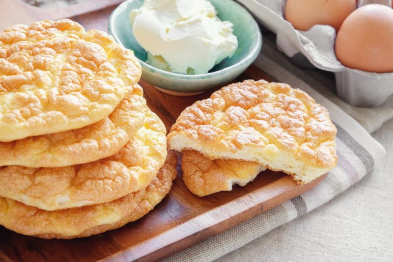 A stack of keto cloud bread, next to a piece of the bread torn in half, a bowl of cream cheese, and some eggs