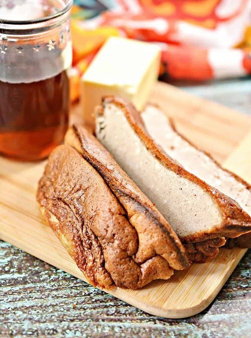 A wooden board with a French bread keto loaf in slices, next to a jar of syrup