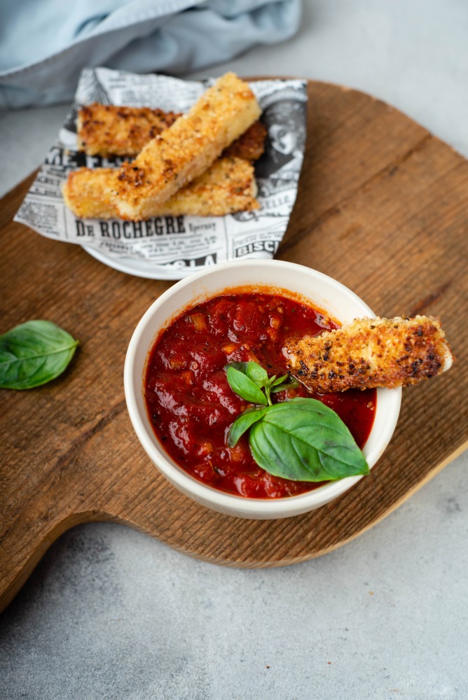 A white bowl with marinara sauce and a fried mozzarella stick, next to some paper with more mozzarella sticks