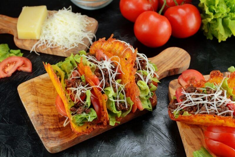 A wooden board with keto tacos that use cheese to create the taco shells, next to the various ingredients, like cheese and tomatoes