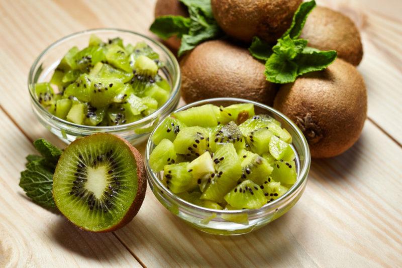 Two bowls of chopped kiwis, a kiwi that has been sliced in half and various whole kiwis